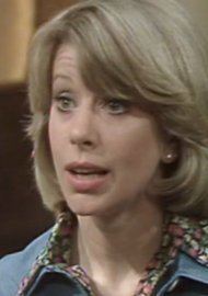 Sheila Fearn talking to someone, with a serious face in a scene from the 1976 TV series, "George and Mildred". She has blonde shoulder-length curled hair and side bangs, wearing stud earrings and a black floral blouse under a blue collared blouse.