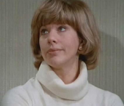 Sheila Fearn looking at something in a snobbish way in a scene from the 1964 film, The Likely Lads. She has short blonde hair with a fringe, wearing stud earrings, and a white knitted mock neck sweater.