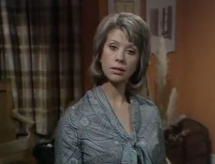 Sheila Fearn looking at something with a snobbish look and blonde shoulder-length curled hair and side bang with an orange wall, wall decors, and a brown checkered curtain in the background, while wearing a printed gray-knotted long sleeve.