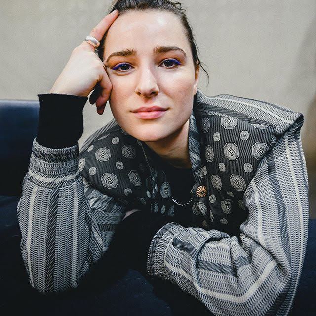 Shelby Flannery pointing at her head while wearing a gray and black long sleeve blouse, ring, and necklace