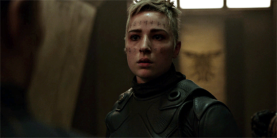 Shelby Flannery with tattoos on her face and blonde short hair while wearing black body armor in a scene from the 2014 television series, The 100
