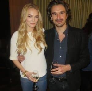 Shelby Welinder and Edward Akrout are smiling while holding a glass of wine. Shelby with long blonde hair and wearing a white blouse while Edward wearing a black coat over a gray shirt and black pants.