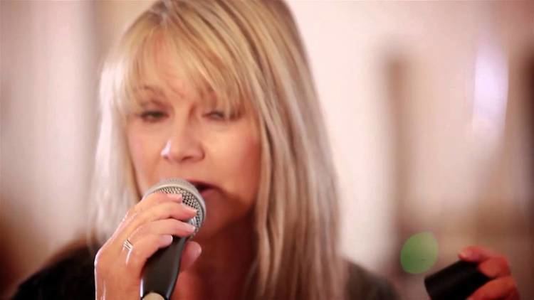 Shelley Preston singing while holding a mic, with blonde hair, and wearing a black shirt.