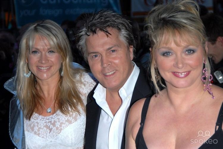 Shelley Preston, Mike Nolan, and Cheryl Baker are smiling. Shelley with blonde hair, wearing earrings, a necklace, and a white lace top, Mike wearing a black coat over white long sleeves, and Cheryl wearing pink earrings and a sleeveless black top.