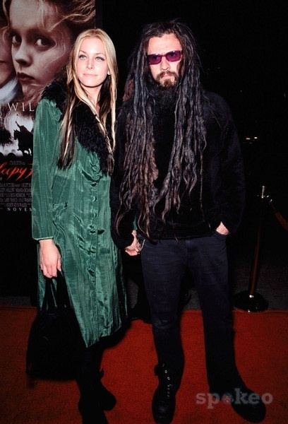 Sheri Moon Zombie smiling while holding hands with Rob Zombie. Sheri with long blonde hair and wearing a green dress with black fur while Rob with long hair, wearing sunglasses, a black long sleeve shirt, and jeans.