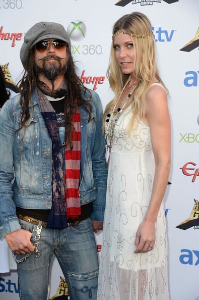 Sheri Moon Zombie smiling with Rob Zombie. Sheri with long blonde hair and wearing a white spaghetti dress while Rob with long hair, wearing a gray hat, sunglasses, a denim jacket, and blue jeans.