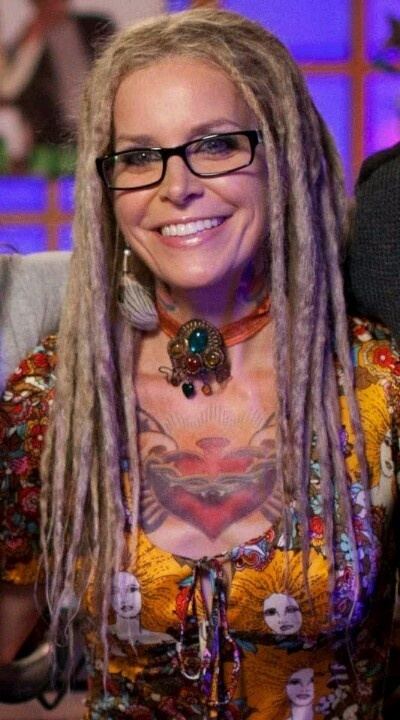 Sheri Moon Zombie smiling with dreadlocks hair, with a tattoo on her chest, wearing eyeglasses, a necklace, and a multi-colored printed shirt.