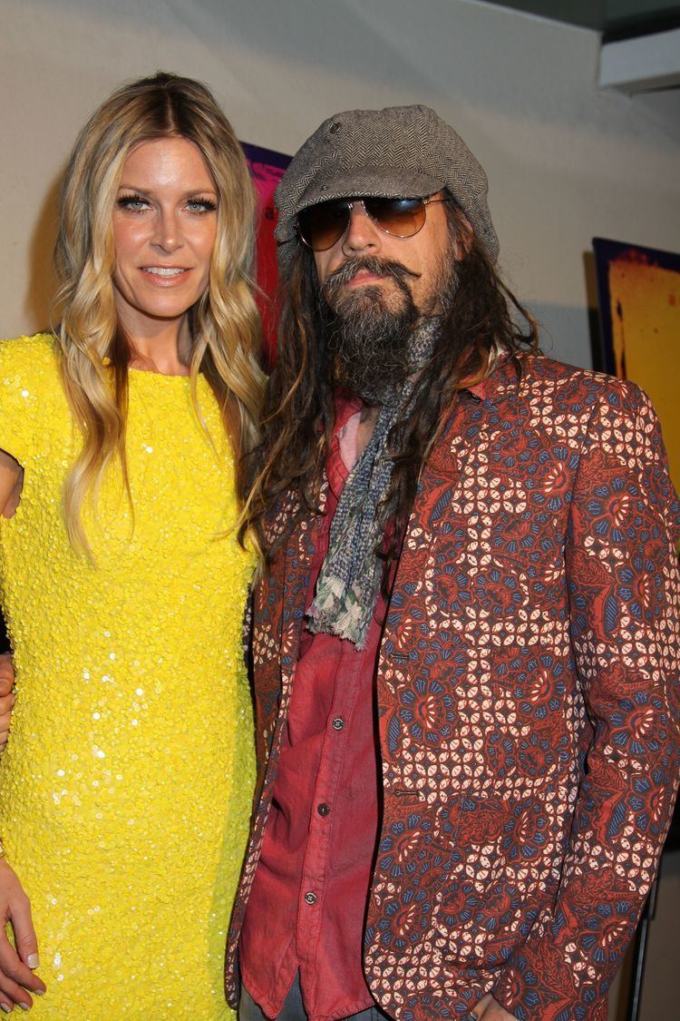 Sheri Moon Zombie smiling with Rob Zombie. Sheri with wavy blonde hair and wearing a yellow dress while Rob with beard and mustache, wearing a gray hat, sunglasses, a multi-colored printed coat over red long sleeves.