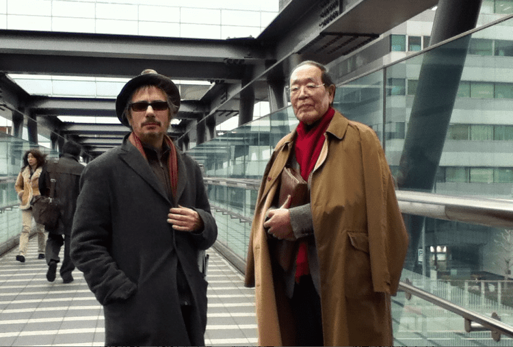 Shigehiko Hasumi wearing eyeglasses, a red scarf, a brown coat, and holding a brown bag with a man wearing a hat, sunglasses, and a black coat.