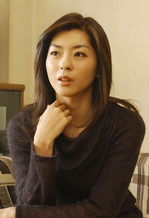 Shinobu Nakayama looking at something while hand on her neck and wearing a black long sleeve blouse
