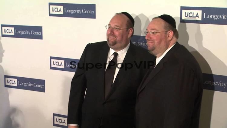Shlomo Rechnitz and Steve Rechnitz are smiling, both are wearing black kippahs, eyeglasses, black coats over white long sleeves, and black neckties.