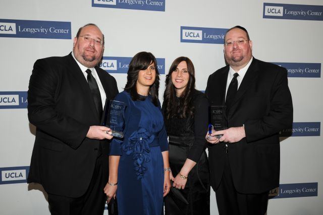 Steve Rechnitz, Steve's wife, Tamar Rechnitz, and Shlomo Rechnitz (from left to right) are smiling while Steve and Shlomo holding plaques and both are wearing eyeglasses, black coats over white long sleeves, and black neckties. Steve's wife is wearing a blue dress while Tamr is wearing a black dress.