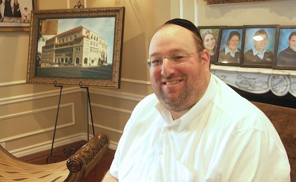 Shlomo Rechnitz smiling in front of a painting and picture frames, with a thin beard and mustache, wearing a black kippah, eyeglasses, and a white polo shirt.