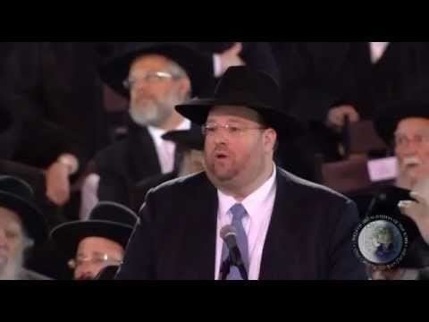 Shlomo Rechnitz is talking in front of many people, wearing a black hat, eyeglasses, a black coat over white long sleeves, and a gray necktie.