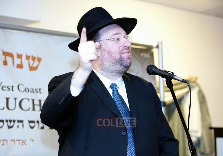 Shlomo Rechnitz is talking in front of a microphone while pointing his finger above, with beard and mustache, wearing eyeglasses, a black hat, a black coat over white long sleeves, and a blue necktie.