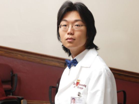 Sho Yano wearing eyeglasses, lab gown, white long sleeves and blue bow tie