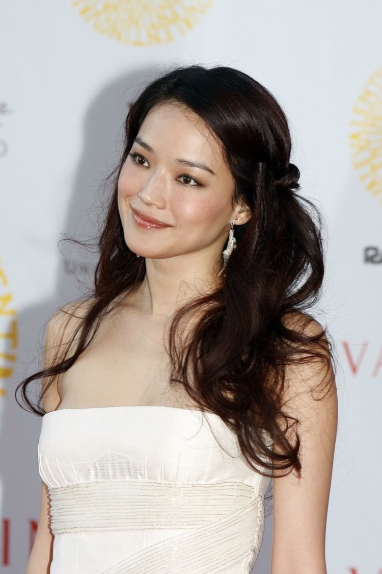 Shu Qi is smiling, has long black hair, and wears silver earrings, and a white off-shoulder top.