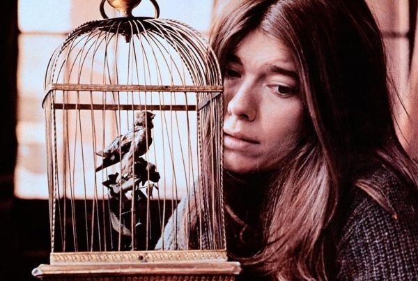 Sian Barbara Allen with a sad face while looking at the bird inside a cage and wearing a black knitted top.