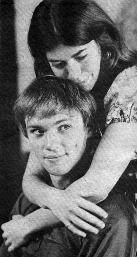 Sian Barbara Allen with a serious face while hugging Richard Thomas with a tight-lipped smile. Sian Barbara with a wavy hair and wearing earrings and a ring on her right pinky while Richard is wearing black long sleeves.