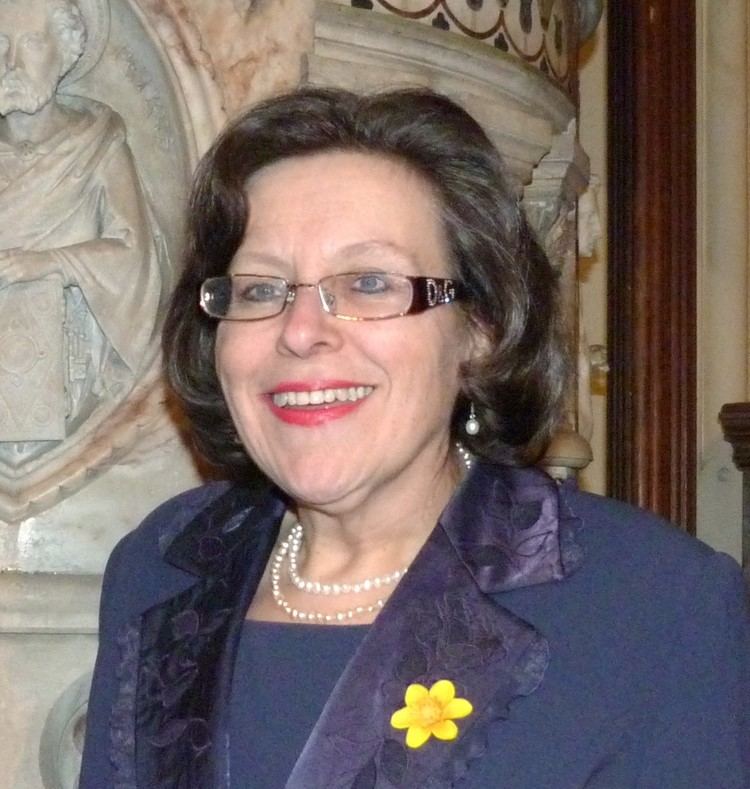 Sian Barbara Allen smiling, with wavy short hair, wearing eyeglasses, pearl earrings, a pearl necklace, and a blue blazer with a yellow flower over a blue top.