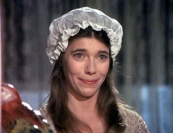 Sian Barbara Allen with a sad face, wavy hair, wearing a white headdress in a scene from The Waltons, a 1972 American historical drama television series. She is wearing and a gray top.