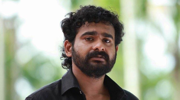 Sidharth Bharathan with a serious face, mustache, and beard while wearing a black polo