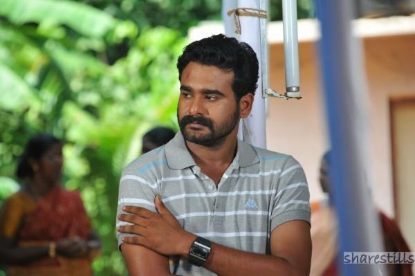 Sidharth Bharathan looking at the left side while holding his arm, with mustache and beard, and wearing a white and gray striped polo shirt and wristwatch