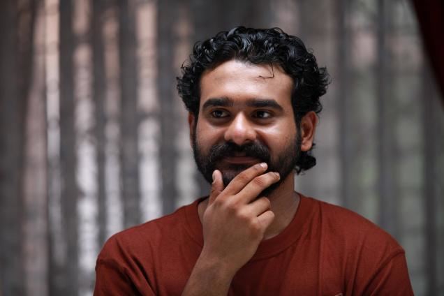 Sidharth Bharathan with a tight-lipped smile, mustache, and beard while hand on his chin and wearing a red t-shirt