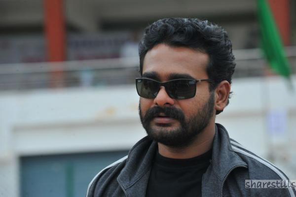 Sidharth Bharathan looking at something, with mustache and beard, while wearing black sunglasses, gray jacket, and black t-shirt