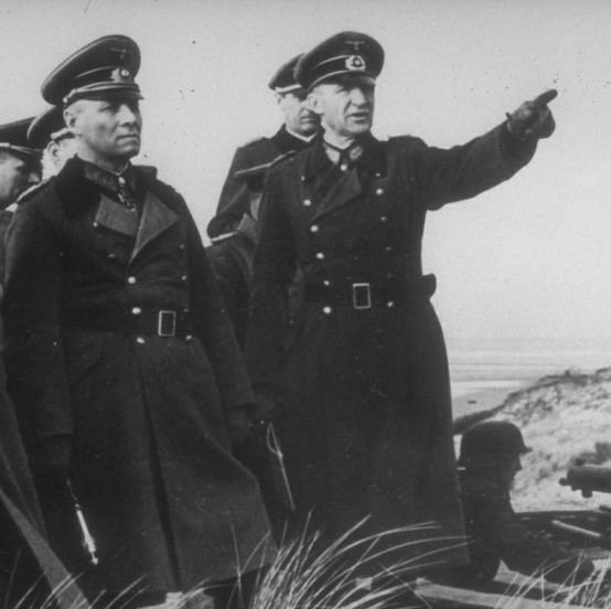 Erwin Rommel (left ) is serious, wearing a service cap, an army service uniform under a black coat with buttons on each side, and a black belt. Siegfried Macholz (right) is serious, right-hand pointing, wearing an army service uniform under a black coat with buttons on each side and a black belt. Behind them are men wearing a service cap and a service uniform. Beside Siegfried (right) is an army person laying down.