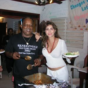 Sigal Erez smiling with the man beside him while holding a plate and wearing a white long sleeve blouse, red necklace, and white pants