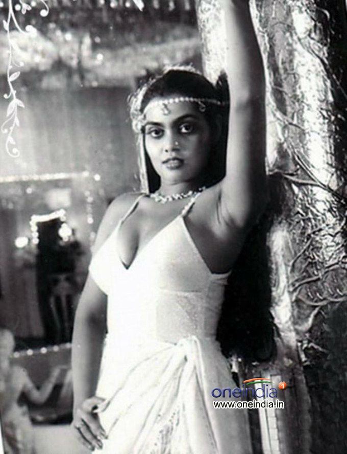 Silk Smitha raising her hand while posing and wearing a sexy sleeveless top, wrap-around skirt, and some pieces of jewelry