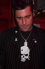 Simon Hobart is serious,  looking down he has black hair wearing a silver dog tag necklace, a black shirt with a white skull printed on it under a black coat with stripe of white line and a skull pins on collar and right chest