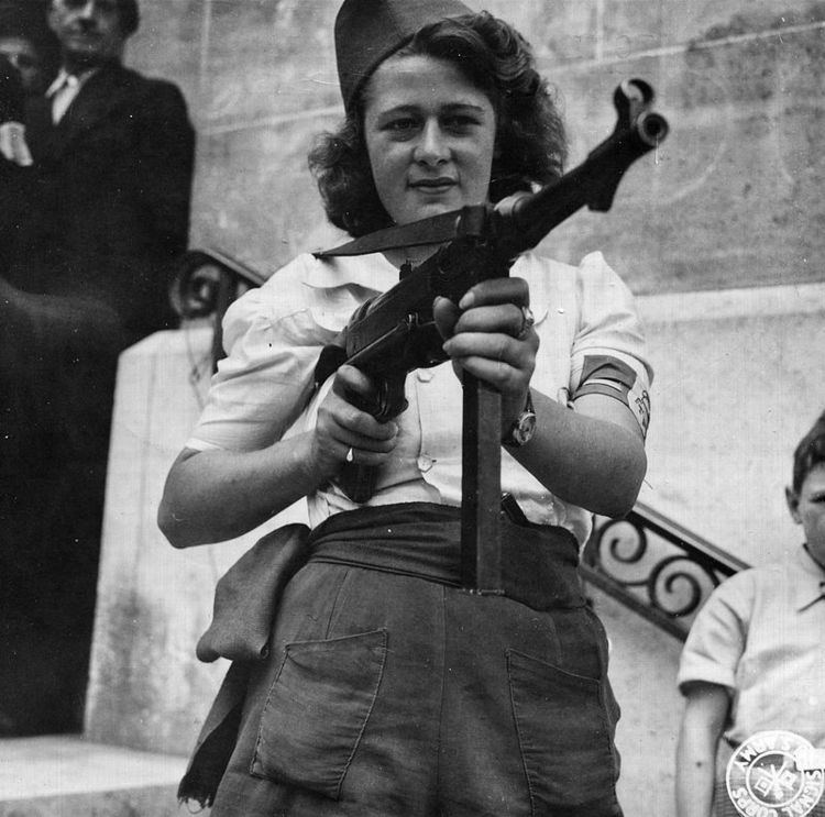Simone Segouin posing with a German MP 40 during the liberation of Chartres while wearing blouse, shorts, wristwatch, hat, and rings