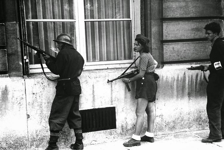 Simone Segouin holding a German MP 40 with two men beside her during the liberation of Paris and she is wearing a blouse, shorts, hat, and shoes