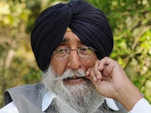 Simranjit Singh looking serious and touching his mustache & having a beard, wearing a white shirt under a gray vest, eyeglasses, and a blue turban