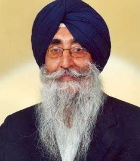 Simranjit Singh looking serious with a beard and a mustache, wearing a white shirt under a blue coat, eyeglasses, and a blue turban
