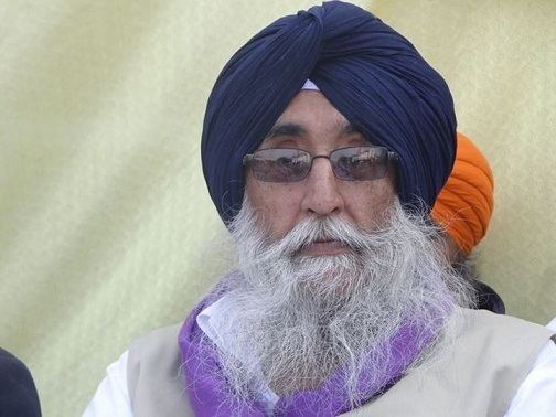 Simranjit Singh looking serious with a beard and a mustache, wearing a white shirt, purple scarf, black eyeglasses, and blue turban