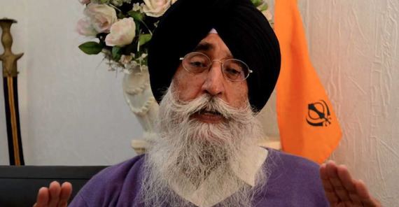 Simranjit Singh speaking having a beard and a mustache with a flower vase, sword, and a Sikh flag in the background, wearing a white shirt under a long purple jacket, eyeglasses, and a black turban