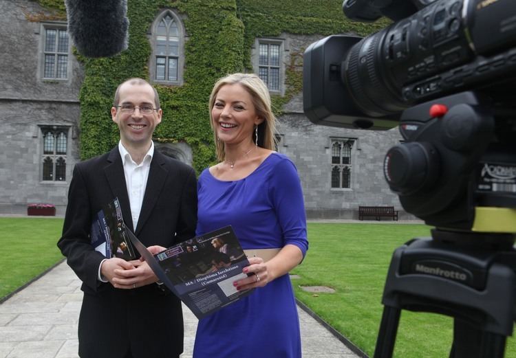 Siún Nic Gearailt with a smiling face and blonde hair, wearing a blue dress, a brown belt while holding a magazine with a man wearing eyeglasses, a black suit, and holding a magazine in front of a camera.