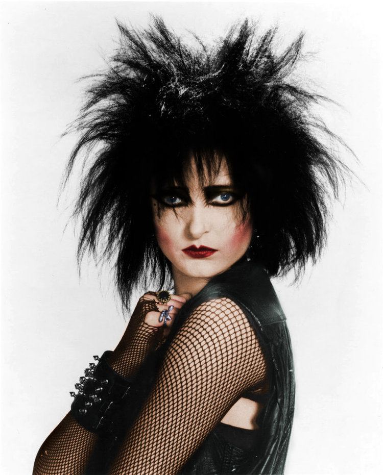 Siouxsie Sioux with messy hair and serious face, wearing a black top and heavy makeup.