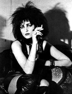 Siouxsie Sioux with messy hair, wearing a black dress while holding a cigarette.