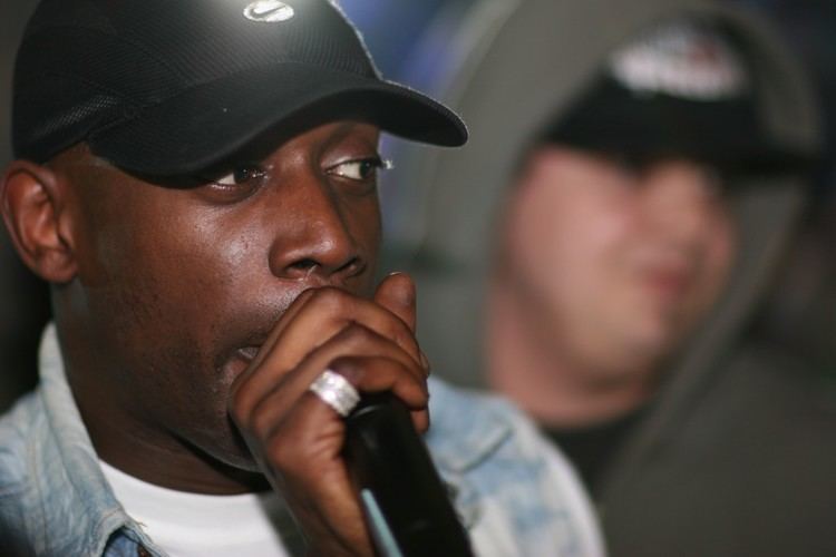 Skibadee looking at something while there's a man on the right side.  Skibadee holding the microphone and wearing a black cap, ring, and white shirt under a blue polo. The man on the right is wearing a black cap and black shirt under a gray hooded jacket