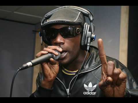 Skibadee singing while holding a microphone, pointing his finger, and wearing a black flat hat, black headphones, a black and yellow shirt under a black Adidas jacket