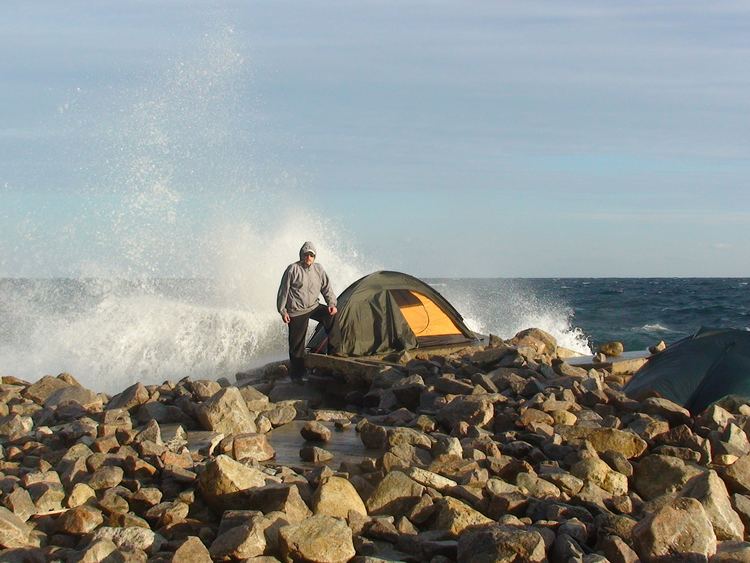 A man is standing beside a green tent surrounded by rocks, wearing a gray hoodie jacket and black pants surrounded by stones on Snake Island also known as Serpent Island belongs Ukraine located in the Black Sea, near the Danube Delta.