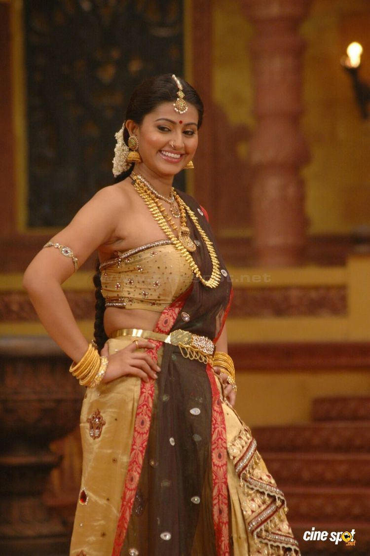 Sneha smiling while hands on her hips and wearing gold, brown, and red Indian dress, earrings, gold necklace, bracelets, armlet, and hair accessories