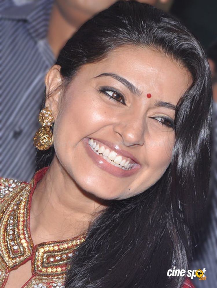 Sneha smiling while wearing gold earrings and a red and yellow blouse with diamonds