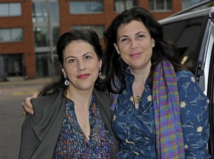 Sofie Allsopp and Kirstie Allsopp are smiling. Sofie wearing earrings and a gray blazer over a multi-colored shirt while Kirstie wearing a necklace and a blue long sleeve top with a multi-colored scarf.