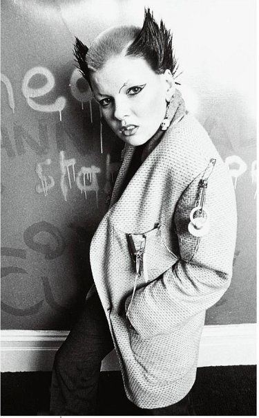 Soo Catwoman is serious while standing while leaning towards the vandalized wall with her right arm, a left hand inside her coat right foot forward, mouth half wide open, has spiky black hair and gray in the middle, funky makeup, wearing skull earrings, scarf and coat with large safety pins.