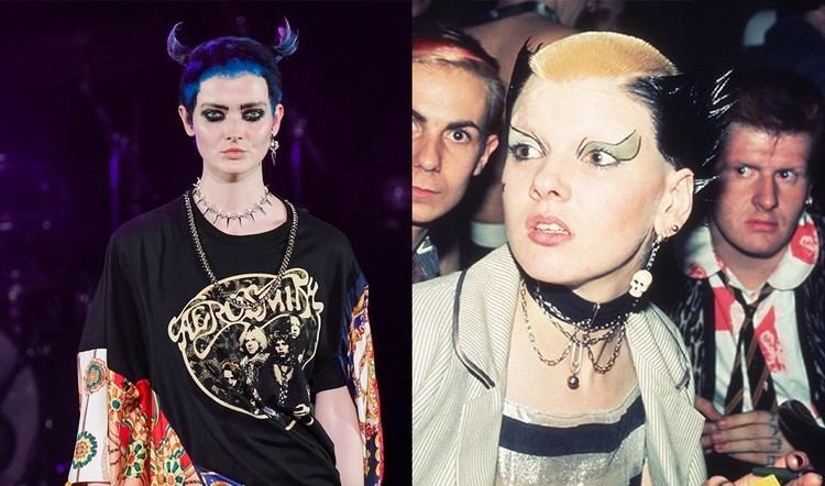 On the left Soo Catwoman is serious, standing, has blue and purple short hair with two horn-like hair on both sides, wearing earrings on her left ears, a gold chain and silver spike necklace, wearing a black aerosmith shirt with flower printed white sleeves. On the right Soo catwoman surprise, looking at her right, holding a pen and yellow paper in her left hand has funky makeup, black spiky hair and blond in the middle wearing skull earrings, choker with chains and bell, black and white striped shirt with white coat, behind her is two men, Simon barker (left) is serious, has red hair and blue shirt, the Marco Pirroni (right) is serious looking at his right, left hand touching the right hand fingers, has black hair, tattoos on fingers,, wearing white polo with red prints, a black and brown striped necktie, animal print jacket.